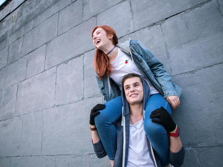 Girl sits on boy's shoulders, in front of a wall. The are having a laugh