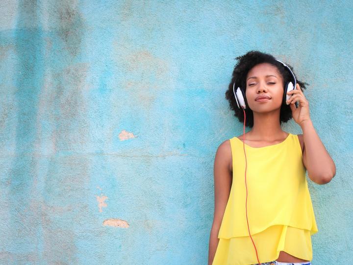 Young girl in yellow dress is standing in front off wall, listening to music